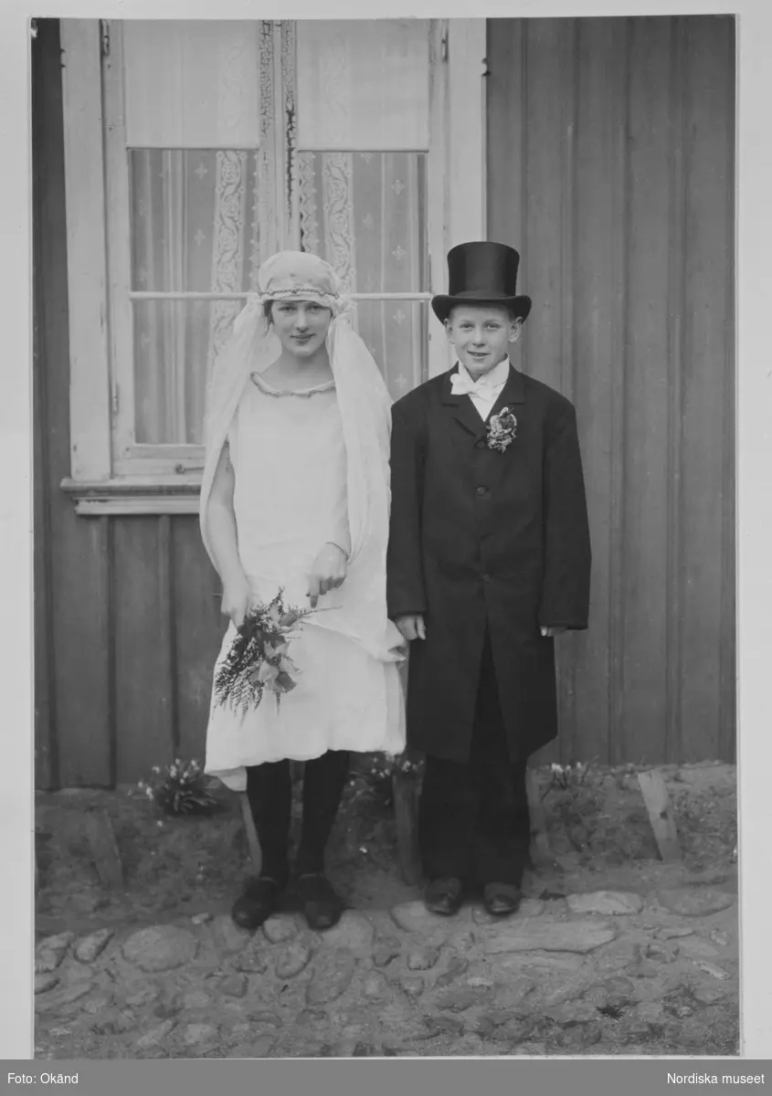 Påskbrud från Lysekil i Bohuslän. En pojke och en flicka klädda för bröllop. Brud och en brudgum står framför ett hus.