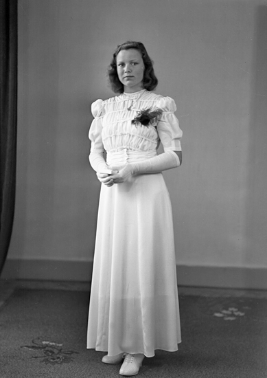 Foto av en kvinnlig konfirmand i lång vit konfirmationsklänning med puffärmar och smock. I händerna håller hon en liten psalmbok.
Helfigur. Ateljéfoto.