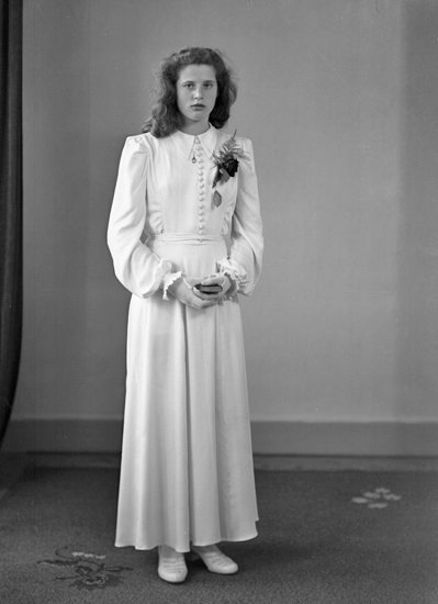 Foto av en kvinnlig konfirmand i lång, vit konfirmationsklänning. 
Helfigur. Ateljéfoto.