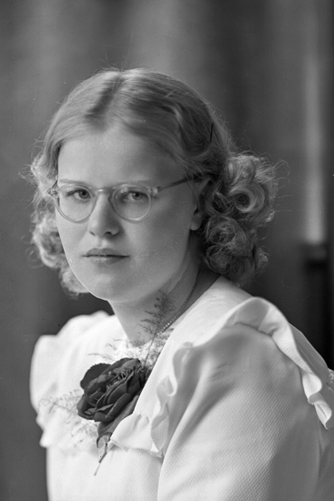 Foto av en ung kvinna i vit konfirmationsklänning (?) och glasögon.
Bröstbild, halvprofil. Ateljéfoto.