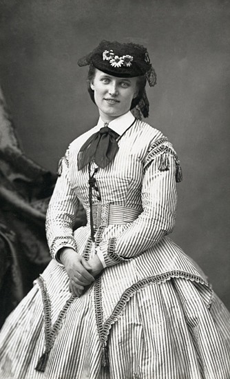 Porträttfoto av Christina Nilsson i Paris. Hon bär randig klänning (krinolin) och en hatt.
Kopia 1920 av äldre foto. (AB).
