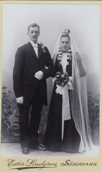 Grupporträtt (helfigur) av ett brudpar, uppklädda i bonjour och svart brudklänning (med vita detaljer.)
Suddigt foto.