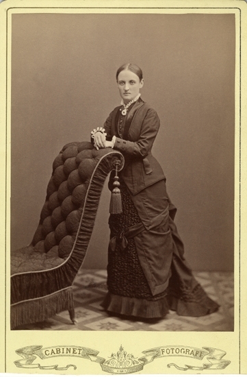 Porträtt (helfigur, ateljé) av en kvinna i mörk klänning med draperad kjol och vit pipkrage. Hon vilar händerna på ryggstödet till en stoppad s.k. halvfåtölj, prydd med fransar och en tofs.