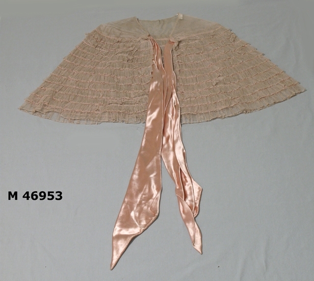 Aftonklänning, rosa satin fleur, ringad rygg, smala axelband, släp. 
Avskuren under bysten. Nedtill en cirka 600 mm klockad del.  
Knäppes med tryckknappar i sidan. 
Längd fram 1600 mm, bak 1900 mm

Tyllcape med band i klänningens tyg.
Capens längd 450 mm. 

Inskrivet i huvudbok  1985