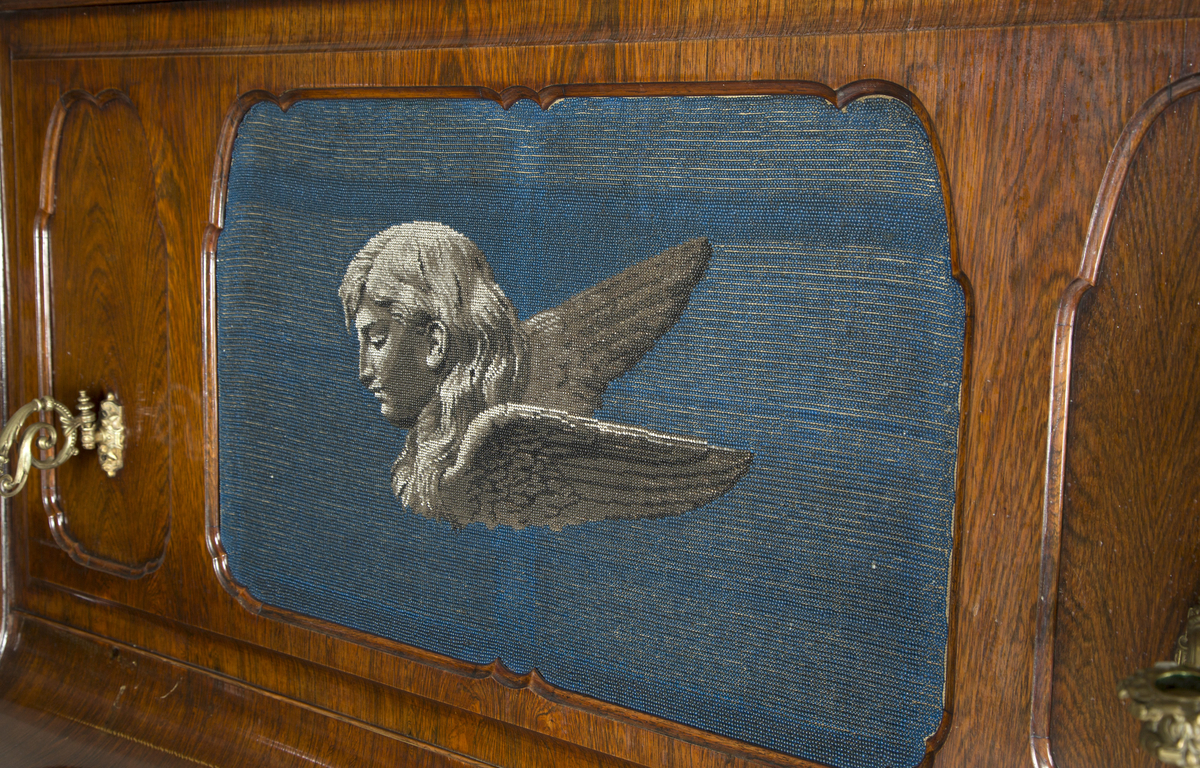 Dekor på frontlokk laget av perler som forestiller et englehode med vinger.