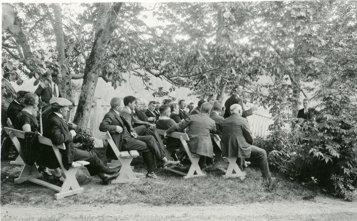 Fra et folkemøte i en park i Amerika. Olaus Islandsmoen taler. Menneskene sitter på trebenker under/ved noen trær.