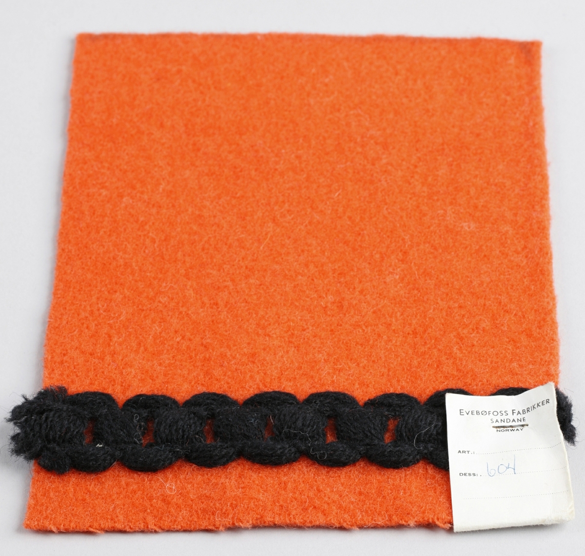 Mønster og stoffprøve for jakke. Rektangel. Orangefarga, vove ullstoff med påsydd kanteband. bandet er trådar sydd saman slik at det liknar forma på ein kjetting.