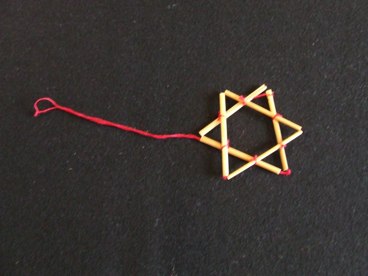 Stjärna av halm. Består av två sammanknutna trianglar. Ögla av rött bomullsgarn. Höjd och bredd 55 mm.