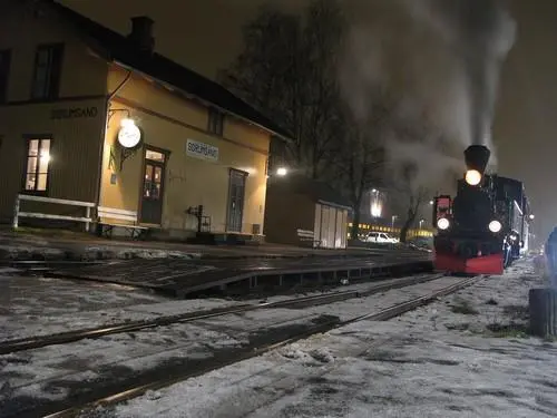 Damplokomotiv foran Sørumsand stasjon en vinterkveld. Det lyser i vinduene på stasjonsmesterkontoret. Snø dekker bakken.