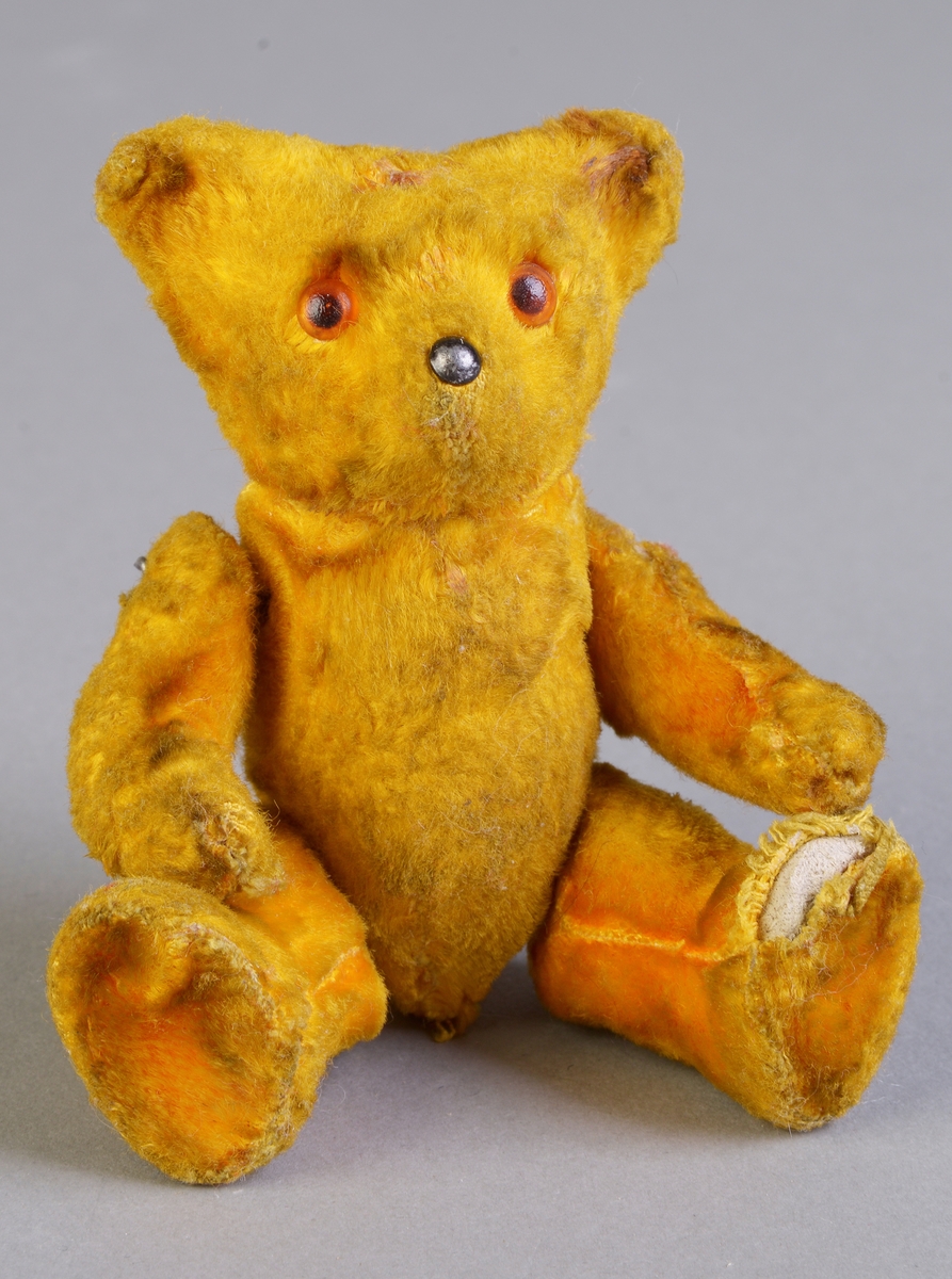 En lien teddybjørn der kroppen muligens er laget av pappmasje. Den er trukket med gult plysjstoff. Armer og bein er festet med ståltråd til kroppen. Øynene er laget av plast og snuten er av metall.
