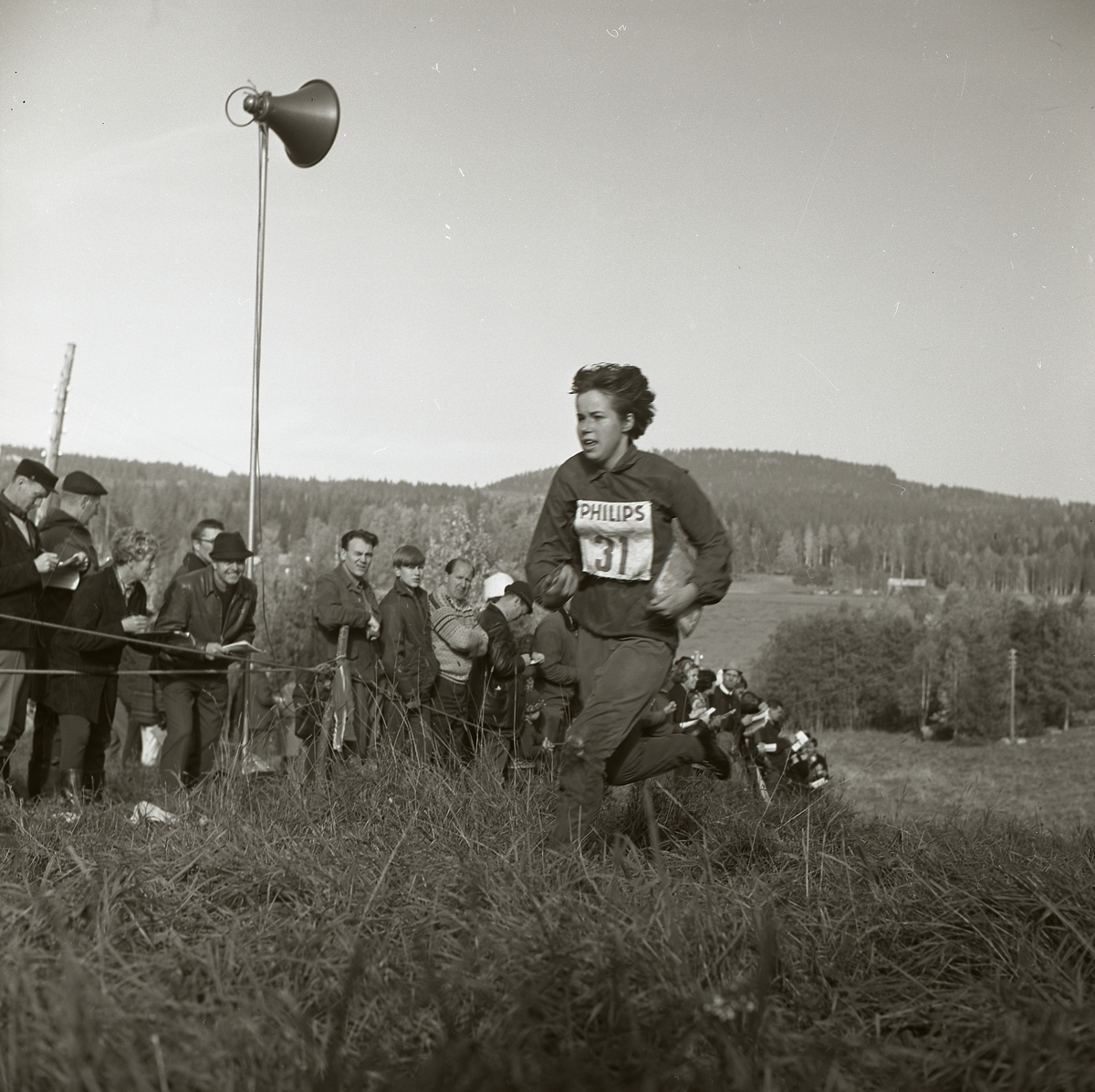 En kvinna springer iförd nummerlapp upp för en backe. Hon bär på en karta och är deltagare i svenska mästerskapen i orientering som hålls i Segersta 1965. Till vänster har en publik samlats bakom en avspärrning. På en stolpe sitter en högtalare som är placerad mitt i folkhopen.