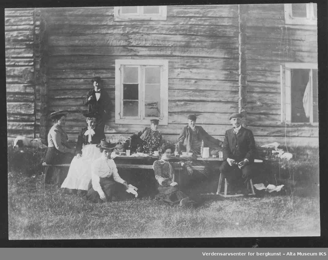 Fire kvinner og fire menn sitter rundt et bord utendørs. Mat og drikke på bordet. Mathisgård.
