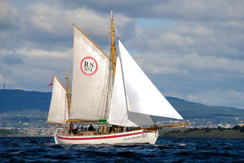 RS1 Colin Archer på vannet med tre seil, land i bakgrunnen.