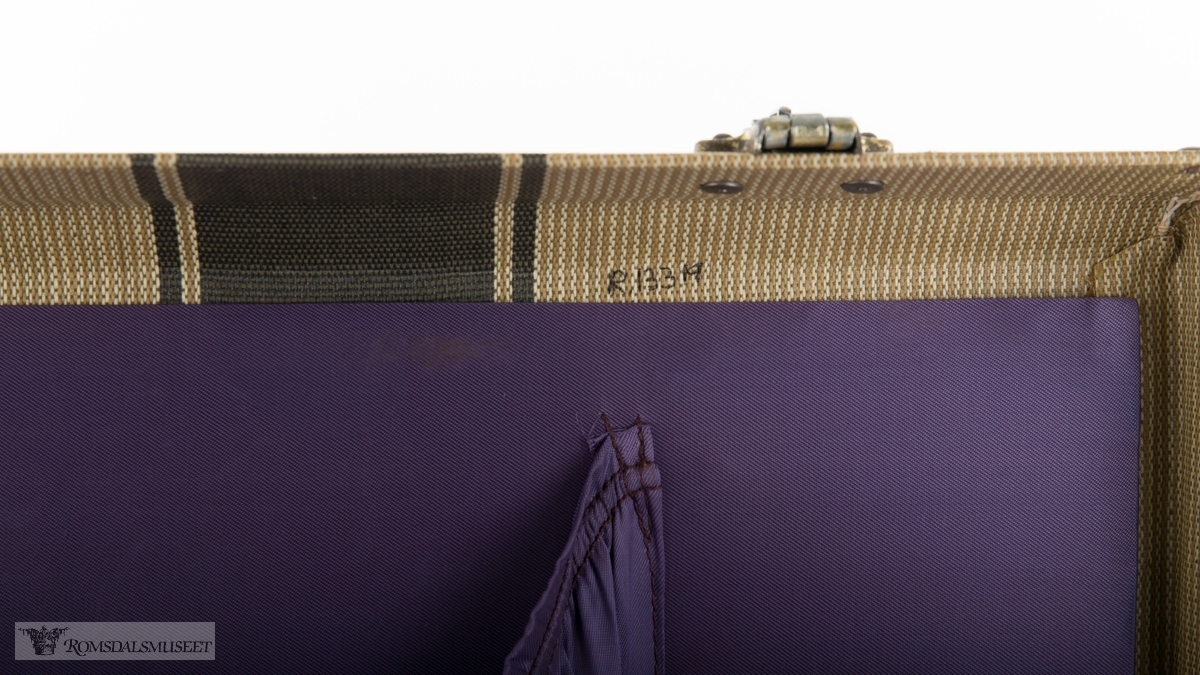 Rektangulær koffert i herdet lerret med forsterkninger av skinn, skinnhåndtak og beslag og lås i messing. Kofferten er kleed med lilla nylonstoff og fire rysjede lommer er sydd inn i trekket på innsiden av lokket og på koffertens sider.