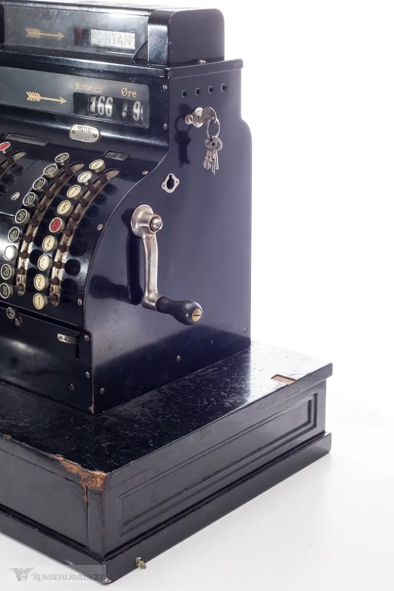 Sortfarget kassaapparat av merke National. Et kontantfelt øverst på apparatet med et felt under for tresifret kronebeløp og tosifret øre. Tre mindre felter over taster merket "KKK", "Kroner" og "Øre".

Fem rader med knapper i front. 
Raden lengst til venstre har bokstaver A-K, samt knapper for "innbetaling", "kreditt" og "utbetaling"
To tasterader på venste side med tall fra 1-9 og 10-90, og tilsvarende på høyre side.
Boks for utskrift av kvittering på vesntre side(?)

Sveiv og nøkler i lås på høyre side og av og på skru på venste side.