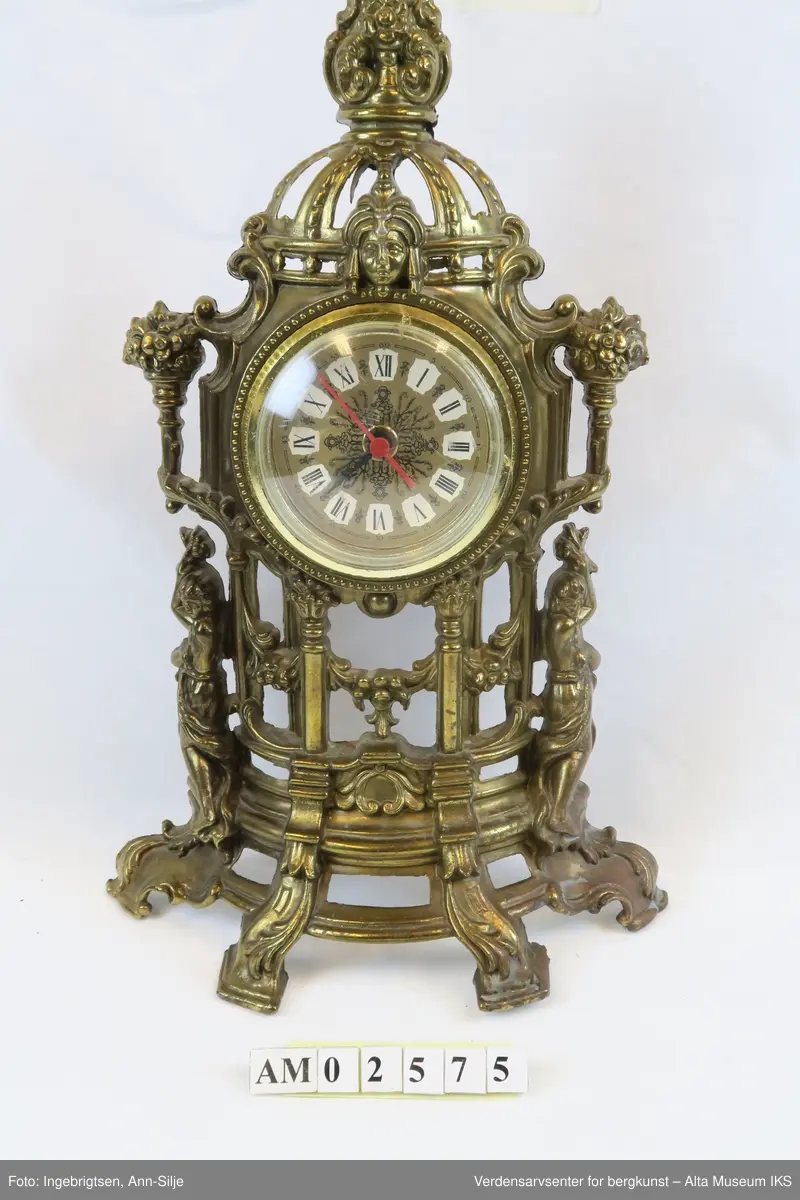 Batteridrevet ur. Stående oppsats med barokk innfatning og innfelt urskive med romertall.