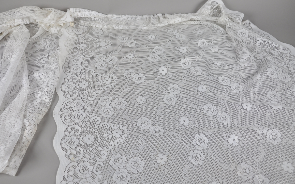 Kniplet gardin av hvit tekstil, med gjennombrutt blomstermønster. Henger i en opphengstråd.