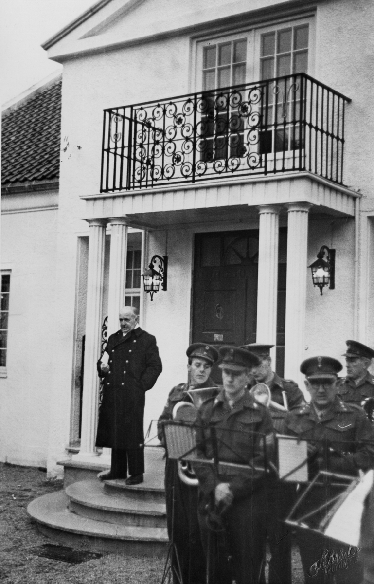 Kommandør Olav Bergersen står på trappen til direktørboligen og holder minnetale over Tordenskiold ved høytidene på Ringve under åpningen av Tordenskiold-museet. I forkant vises et korps.
