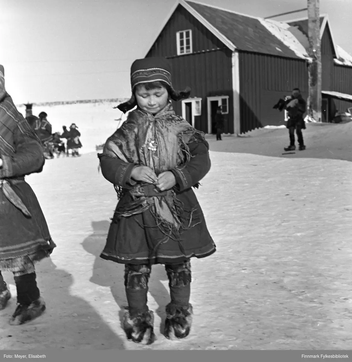 Berit Nilsdatter Logje på plassen utenfor Kautokeino Skoleinternat. Berit har på seg samisk kofte, sjal, sølje, lue, skinnbukser og skaller med skalleband. Det er stor aktivitet på plassen, barn i lek og voksne i arbeid. Sannsynligvis fotografert ved påsketider 1940.