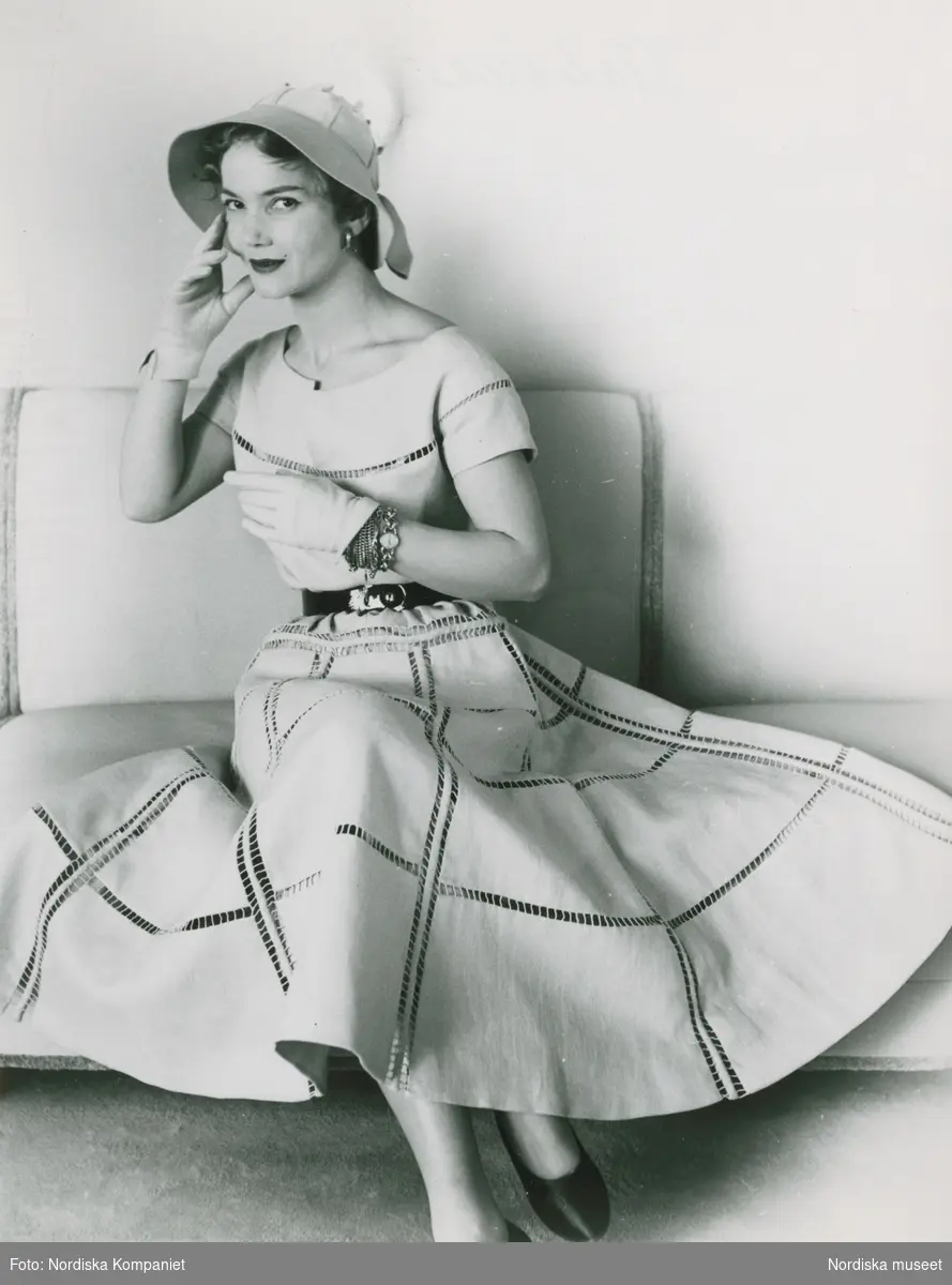 Modell i klänning, hatt, handskar och armband i en soffa. "Italiana".