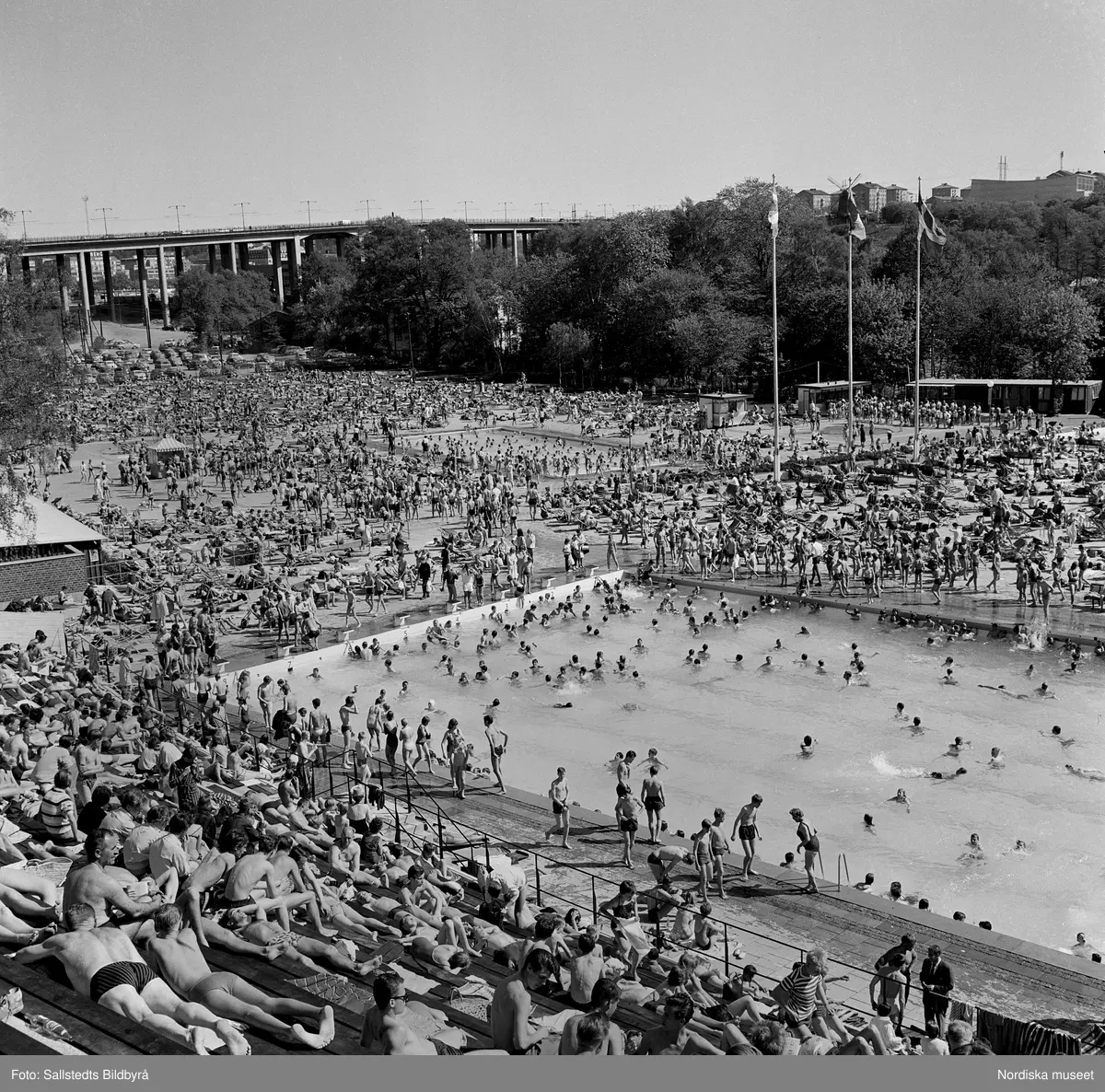 ”Eriksdalsbadet Stockholm” Fullt med badande på Eriksdalsbadet. Stor utomhusbassäng, Skanstullsbron i bakgrunden, Stockholm.