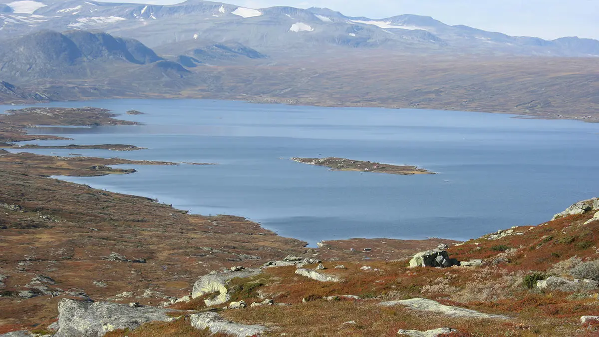 Innsjøen Vinstre sett fra Dragarskaret, i forgrunnen steiner og brunt gras, fjell med litt snø i bakgrunnen.