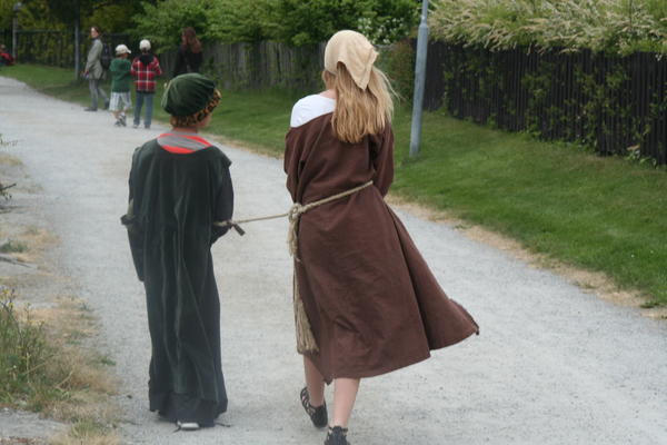 To barn i middelalderklær går med ryggen mot fotografen langs en grusveg.