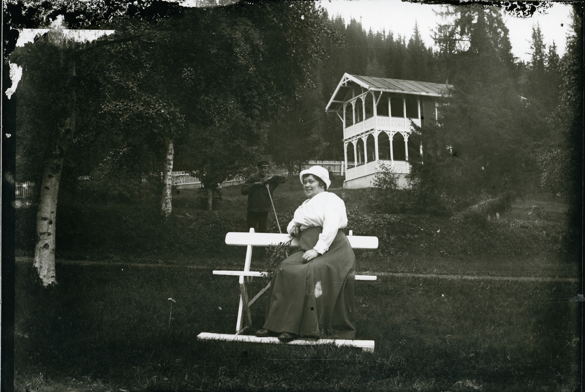 Ei kvinne sit på ein benk ute i ein hage. Bak henne står ein mann med ei rive, ca 1910-1915.