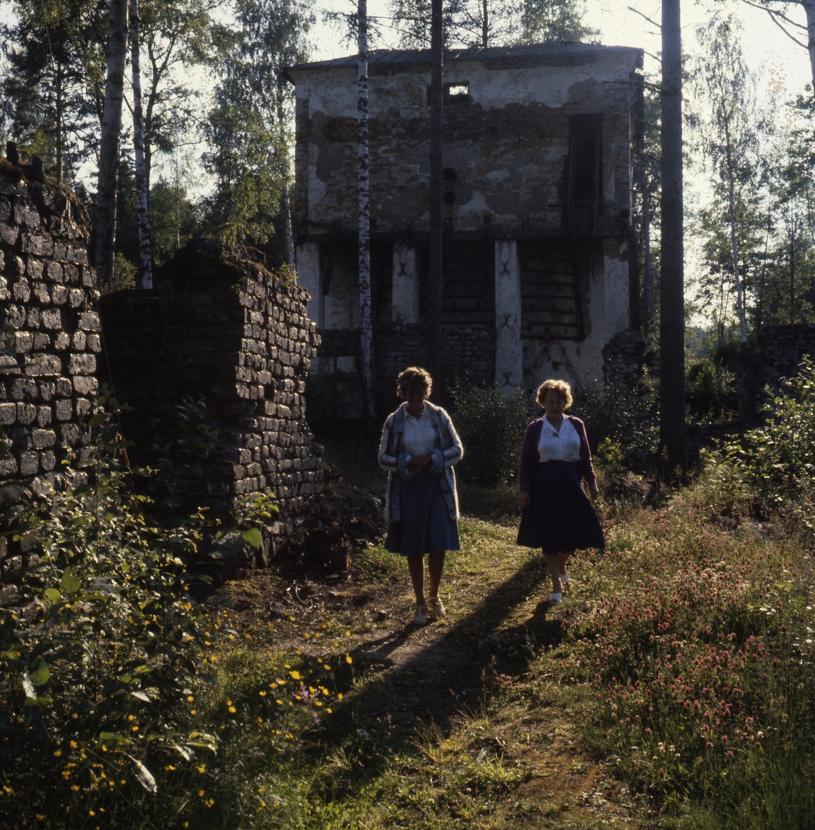 Adéle (till höger i bild och en kvinna till, går framför en raserad byggnad. Det ser ut som mur av slaggsten bredvid dem.