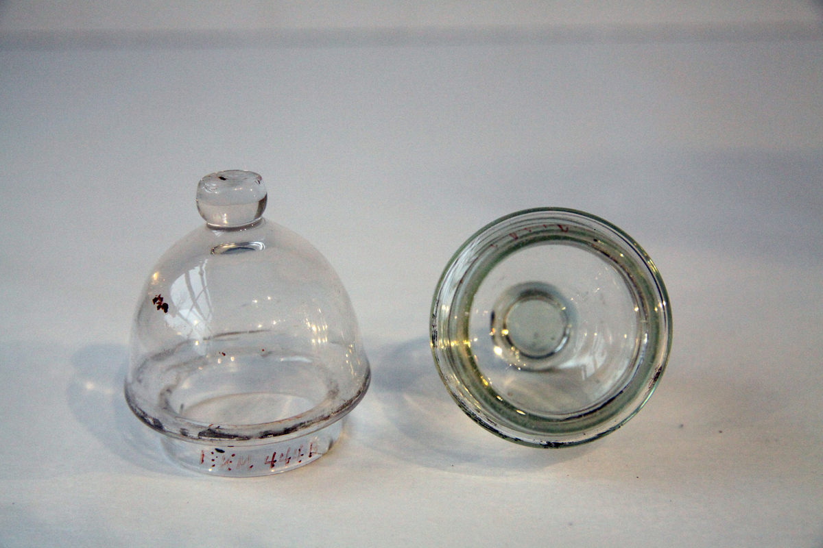 Glassene (b) og (c) har en knopp på toppen, og glass (a) har ei kule av grå gummi på toppen.
