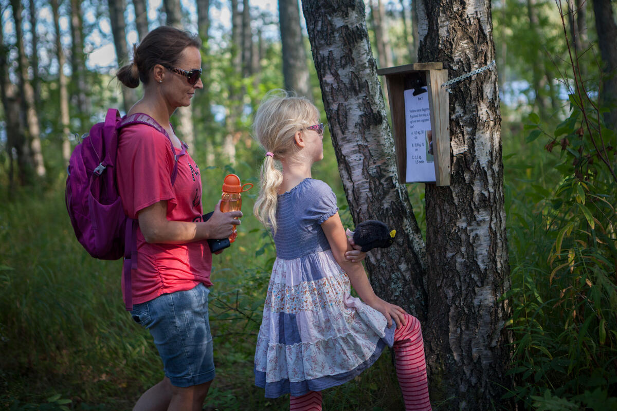 Mor og datter leser på en naturstipost på Blikomøya. Mor har olabukse og t-skjorte på seg og datteren har sommerkjole
