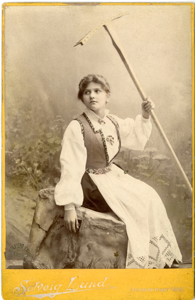 Kolorert studiofotografi av kvinne med Hardangerdrakt sittende med rive i hånda mot blomsterbakgrunn