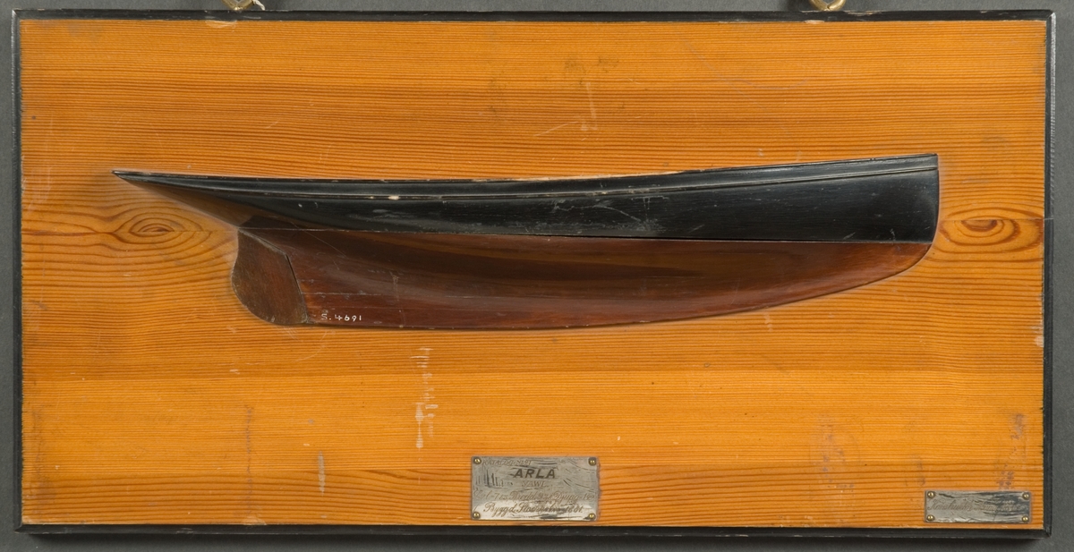 Halvmodell av yawlen Arla, i block av trä, visanda styrbords sida. Skrovet är svar och brunt, polerat. Monterat på polerad ljus träplatta med svart kant. Två silverplåtar med graverad text.