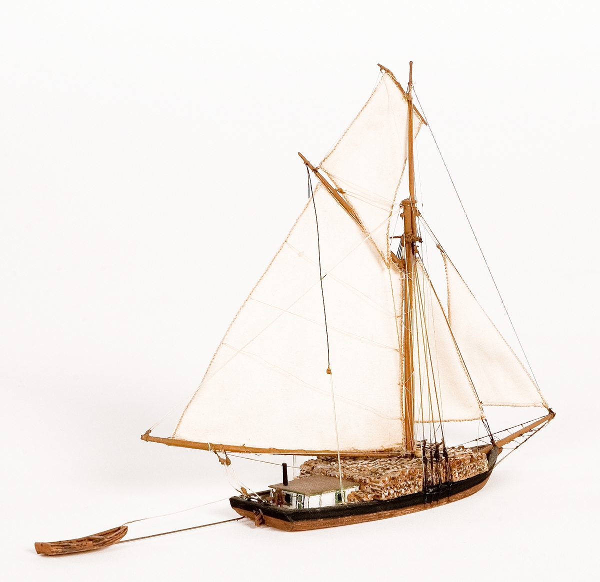 Fartygsmodell på vattenlinje. Roslagsslup, hel i block av trä med mast och segel av bomull. Svarta brädgångar, brunt skrov. Liten båt på släp.Föremålets form: Vattenlinjemodell