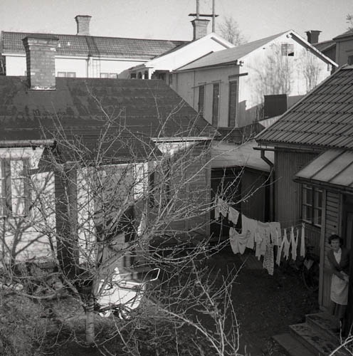 På innergård i Hudiksvall 1954 står en kvinna på trappen och kikar ut. Vardagens göromål som tvättning och barnpassning är representerade via kläder som hänger på tork och en barnvagn ute på gården. I förgrunden skymmer en trädkrona delvis insikten på innergården men bakom denna breder en stadsmiljö med hustak och skorstenar ut sig.