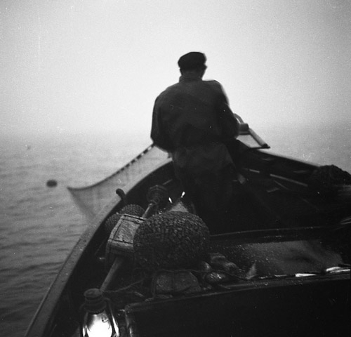 Sommaren 1952 i Skärså tar en fiskare upp skötar ur vattnet till sin båt. Han sitter i fören med resterande fiskeutrustning bakom sig; flöten, nät och en fotogenlampa som sprider lite ljus.