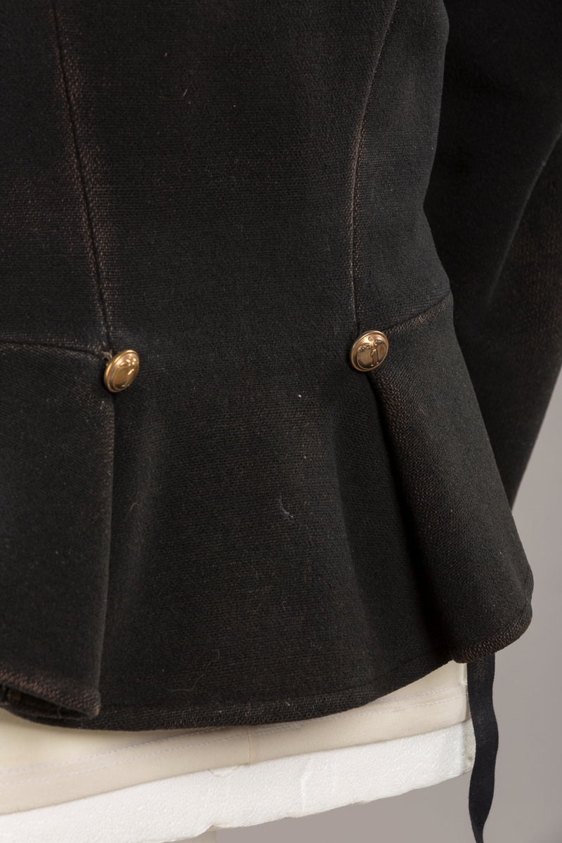 Form: Enkeltspent.

Foret med lin. Forsterket med silke på et lite stykke på ryggen. 12 messingknapper med mønster og tallet 1. Hempe i lin. Bendelbånd med knappehull.