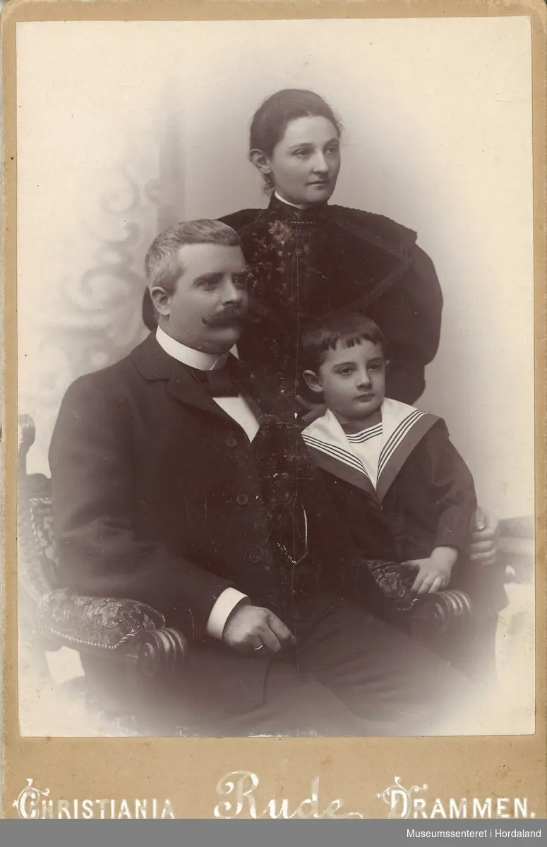 familieportrett av mann, kvinne og gut i finkle, dress med stivesnipp, kåpe og kjole med vide slag, matrosdress
