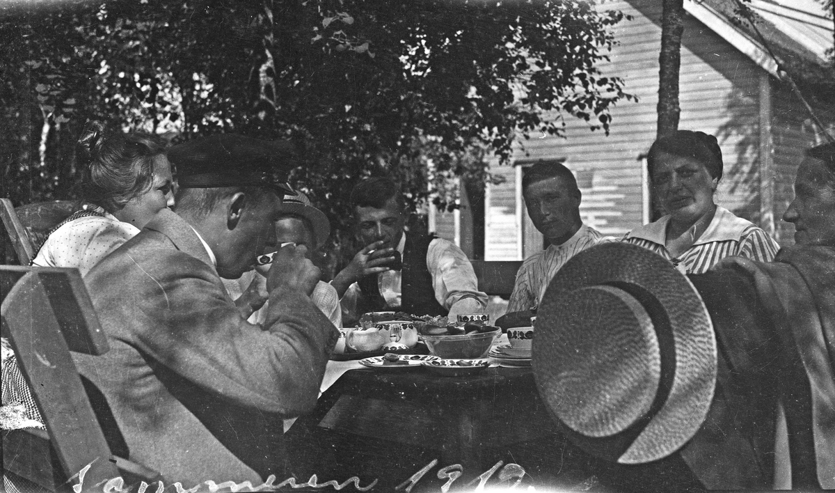 Gruppebilde. Sommer. Tre kvinner, tre menn og et barn slapper av koser seg med mat og drikke i solskinnet. . Hus i bakgrunnen. Hverdagsfoto.