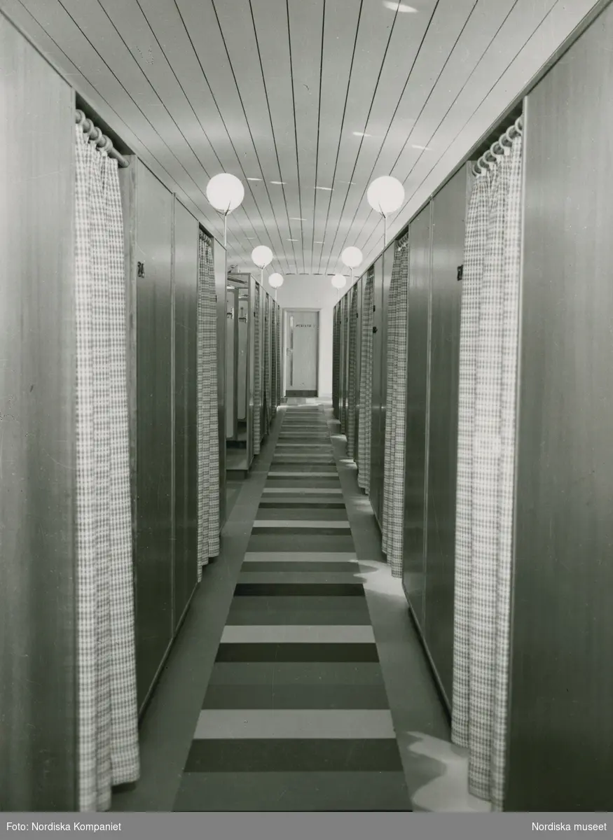 Interiör. Lång korridor med tvärrandig matta. Längs båda sidor bås med rutiga förhängen. Nordiska Kompaniet. Text med blyerts på baksidan: "Hygieniska avdelningen".