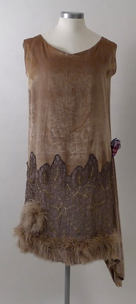 Klänning av ljusgrå silkesammet. På kjolen silverspets och längst ner grå plymer. I midjan en bukett tygblommor i rosa och blått. 1920-talet