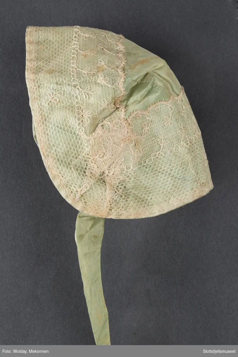 Dåpslue: av samme stoff som capen, sydd av et rett midtstykke og 2 avrundede sidestykker. Pyntet med bred silkeblonde, ecrufarget. Fóret m. Rødbrun blomstret mugg. Ett grønt silkeband, det andre mangler.