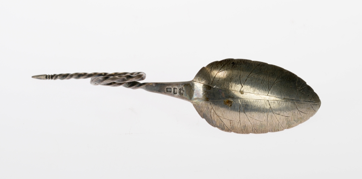 En sukkerskje av sølv. Bladet har hakk i sidene som minner om et bjørkeblad, og på undersiden er det gravert inn nerver i bladet. Skaftet er først flatt før det er snodd/tvunnet. På midten er det en løkke som fungerer som håndtak. Skaftet smalner og i enden smalner det til en spiss. På undersiden er det tre stempel. det første er en blomst som er bymerke for Trondheim. Deretter er det et monogram som viser at det er J.A. Møller som trolig har laget det. Deretter står det 13 1/2 som angir sølvgehalten. På oversiden er det gravert inn en dato.