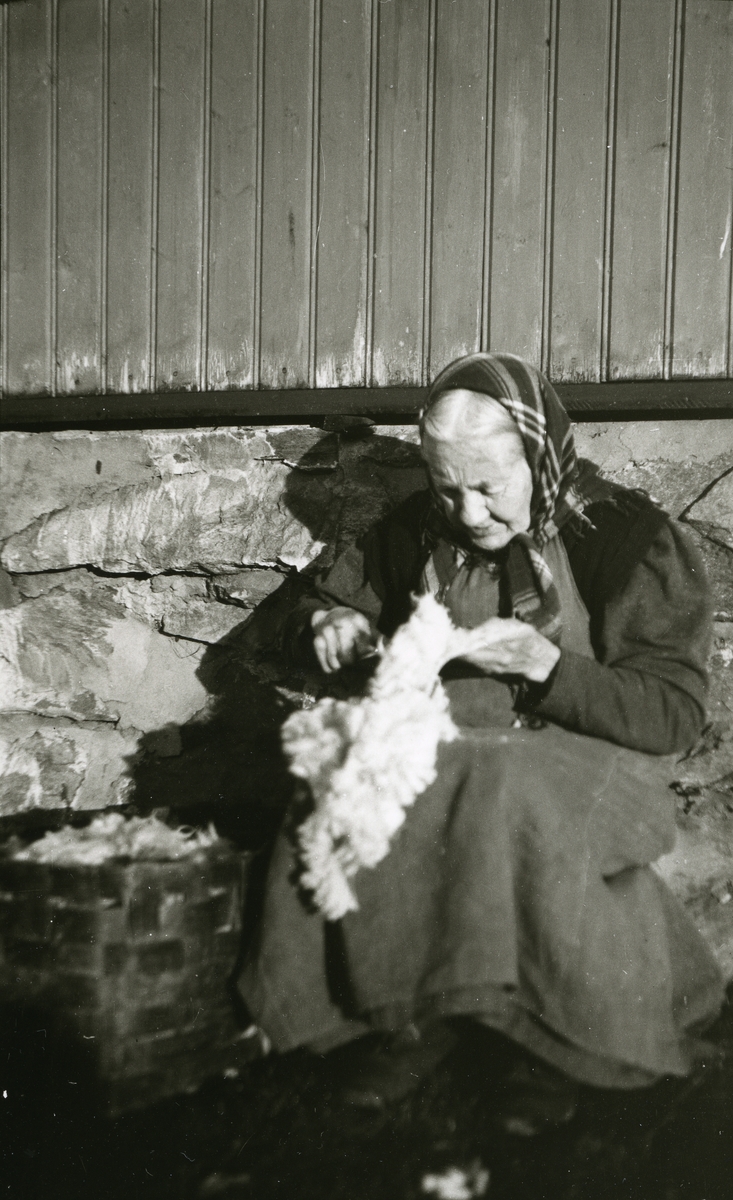 Kvinne sitter innved husvegg (grunnmur) og klipper i skinn i Galåsen, Jons.