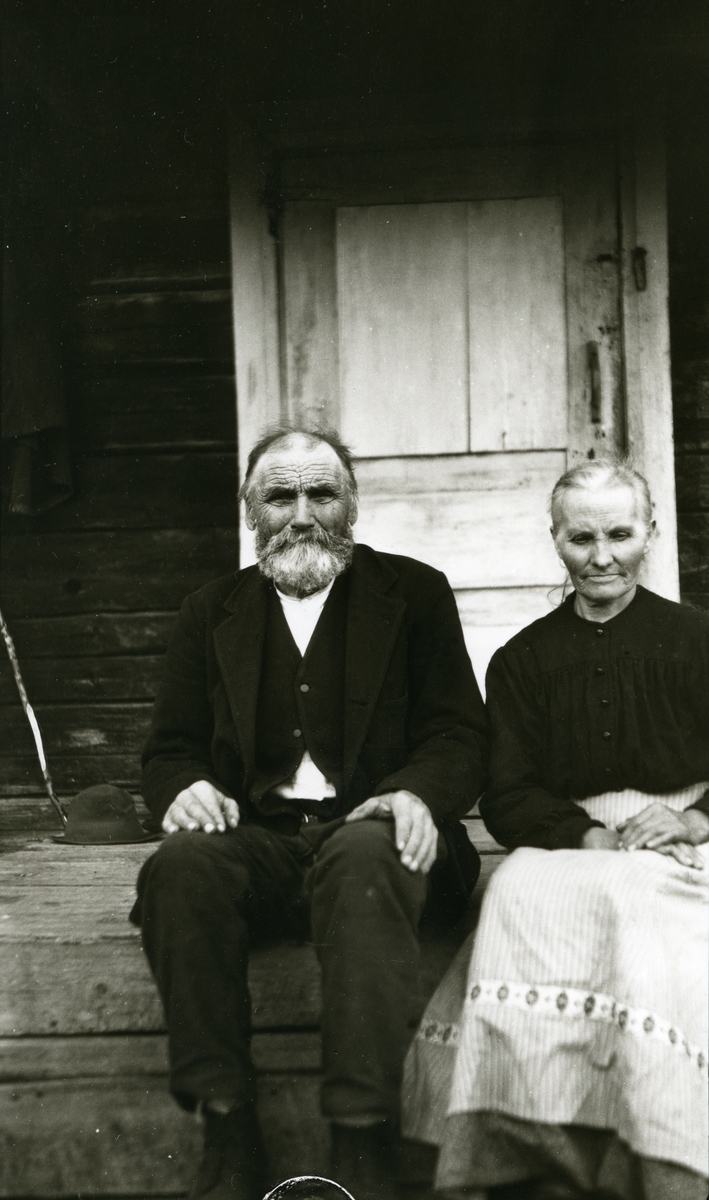 Eldre ektepar sittende ute på trappa (inngangsdøra i bakgrunnen). I Horrmund, Dalarne, Sverige. 
Oluf Karinusen Brändholm (1846-1937) och Maria Jonsdotter (1858-1933) på gården Nya Lövnäs, Horrmund, Transtrand