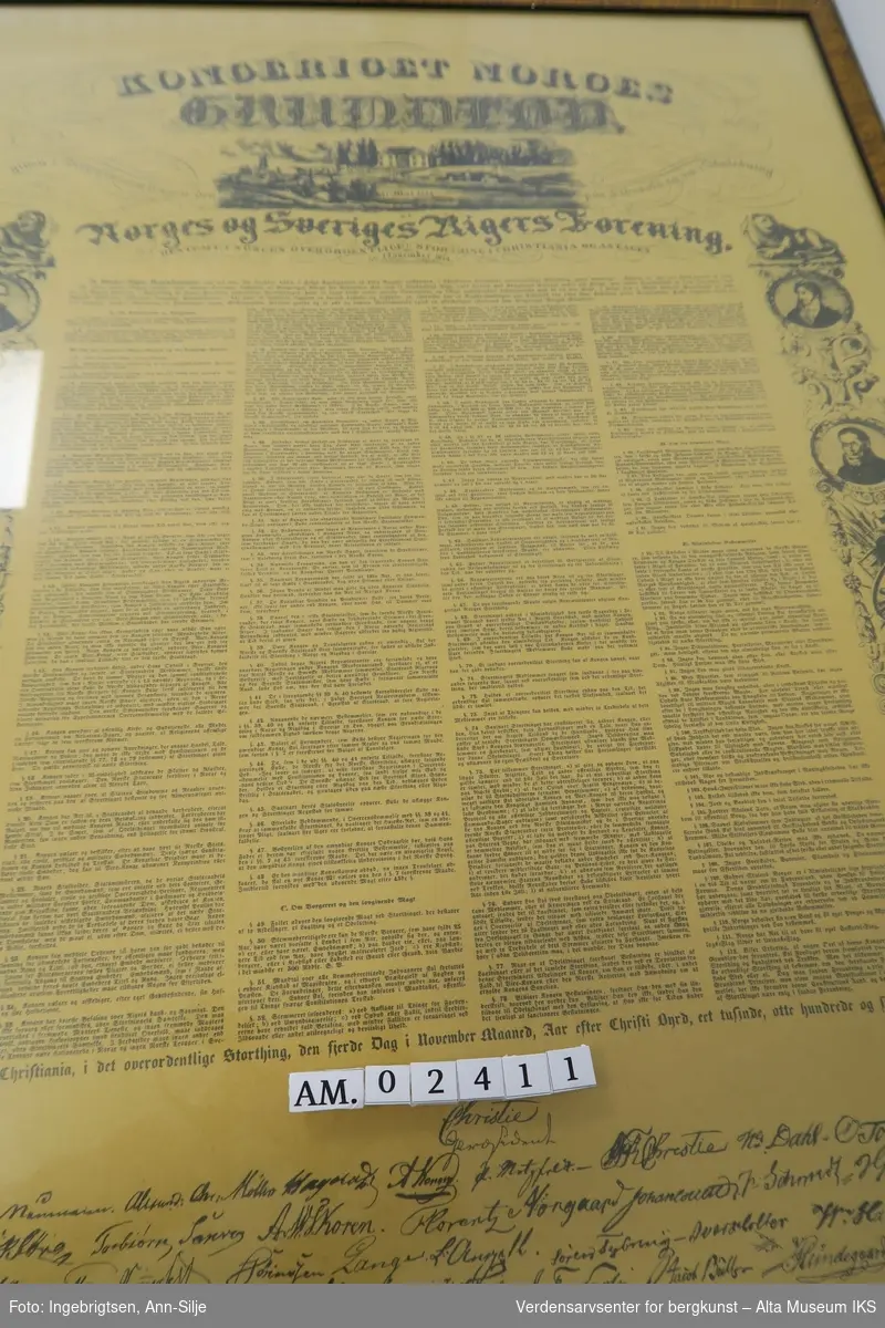Innmrammet "Norges Grunnlov" 1814 med glass og brun ramme. Plakat av grunnloven fra mai 1814 med forandringene fra november samme året. Dekor med bilde av Eidsvoldmenn, engler, løver og blomster.