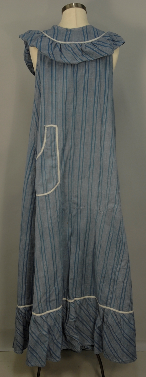 Heldekkende forkle i bomull uten ermer. Det er laget av et blågrått stoff med landsgående striper i en mørkere blå farge. Rundt halsen har det en påsydd rynket kappe i samme stoff, stoffet er skråstilt, det samme sees nederst på forkleet. Forkleet har en lomme på forsiden og åpning bak som kan lukkes med tre lerretsknapper. det er påsydd et kvitt pyntebånd over begge skråkappene og rundt lommen. På forsiden er en rift som det er bøtet på. På forkleets venstre side er et knytebånd i samme stoff som forkleet, dette mangler på høyre side.