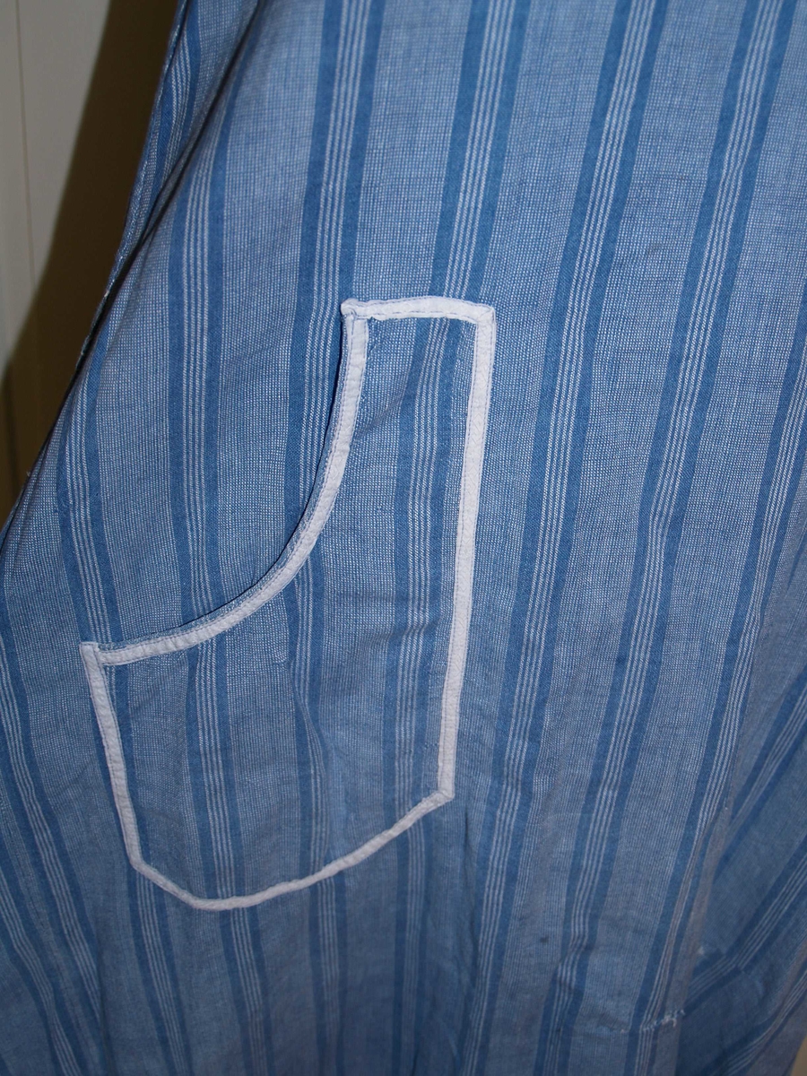 Heldekkende forkle i bomull uten ermer. Det er laget av et blågrått stoff med landsgående striper i en mørkere blå farge. Rundt halsen har det en påsydd rynket kappe i samme stoff, stoffet er skråstilt, det samme sees nederst på forkleet. Forkleet har en lomme på forsiden og åpning bak som kan lukkes med tre lerretsknapper. det er påsydd et kvitt pyntebånd over begge skråkappene og rundt lommen. På forsiden er en rift som det er bøtet på. På forkleets venstre side er et knytebånd i samme stoff som forkleet, dette mangler på høyre side.