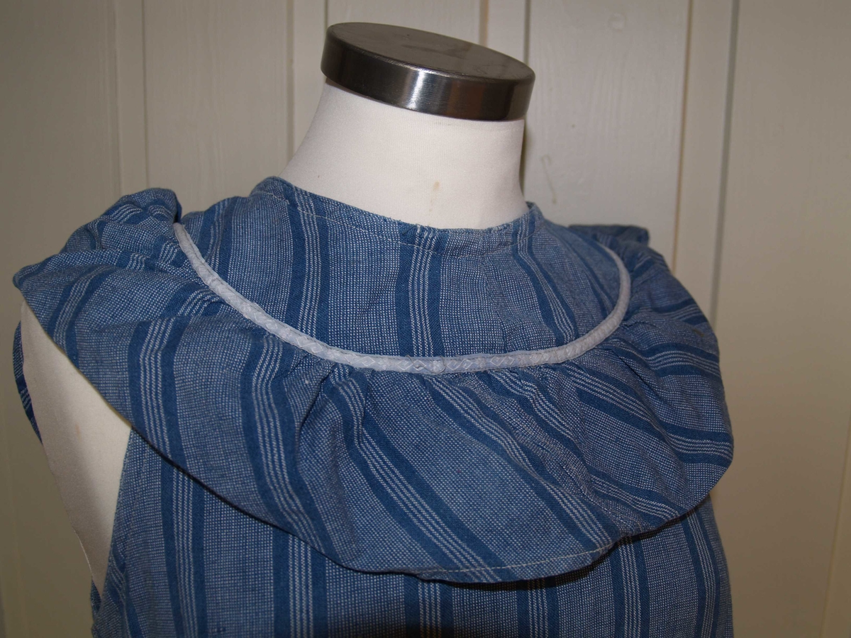 Heldekkende forkle i bomull uten ermer. Det er laget av et blågrått stoff med landsgående striper i en mørkere blå farge. Rundt halsen har det en påsydd rynket kappe i samme stoff, stoffet er skråstilt, det samme sees nederst på forkleet. Forkleet har en lomme på forsiden og åpning bak som kan lukkes med tre lerretsknapper. det er påsydd et kvitt pyntebånd over begge skråkappene og rundt lommen. På forsiden er en rift som det er bøtet på. På forkleets venstre side er et knytebånd i samme stoff som forkleet, dette mangler på høyre side.