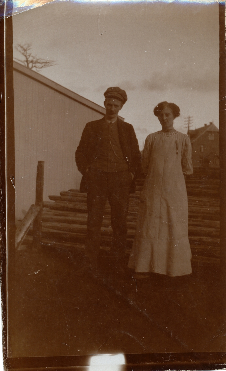 Mann og kvinne foran stabel med tømmer, ant. butikkmedarbeidere hos Meyer. Bygning bak.